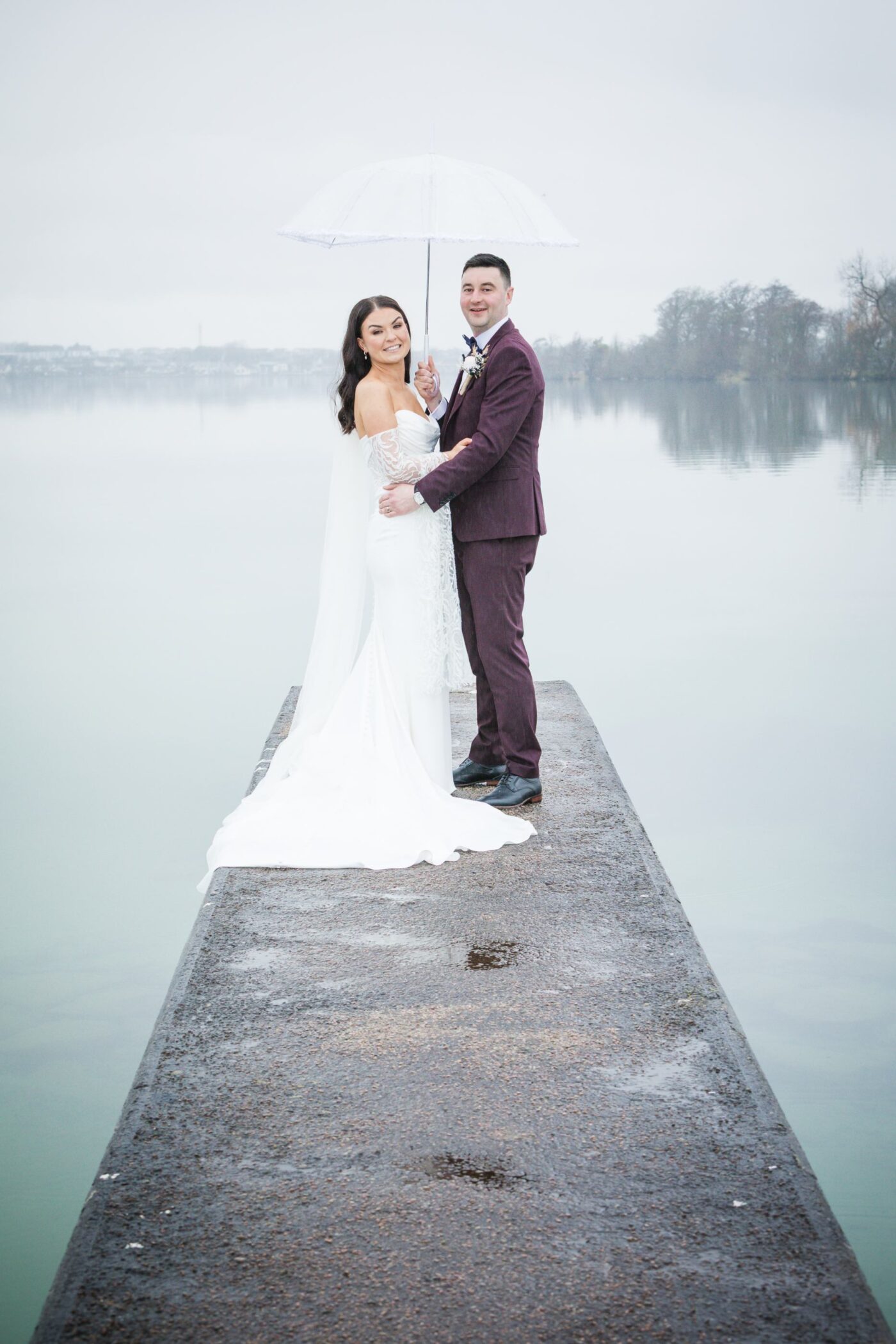 Bride and groom embracing beneath a clear umbrella on a pier overlooking a misty Loughrea Lake.