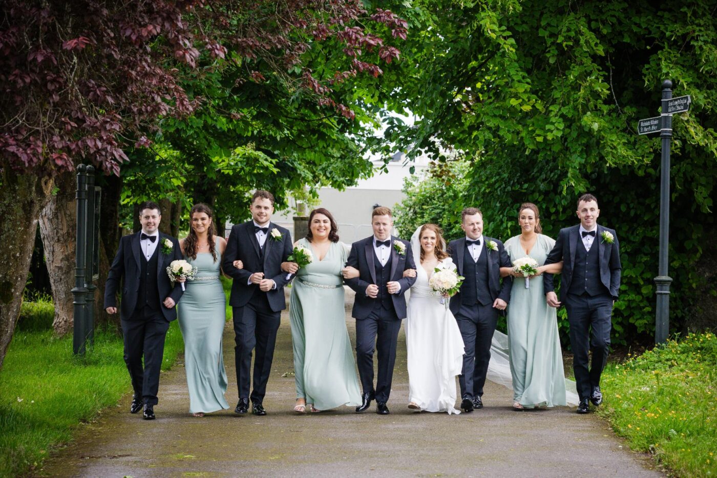 Bride and groom walking arm in arm with bridesmaids in sage green and groomsmen in dark suits along a leafy path in Loughrea.