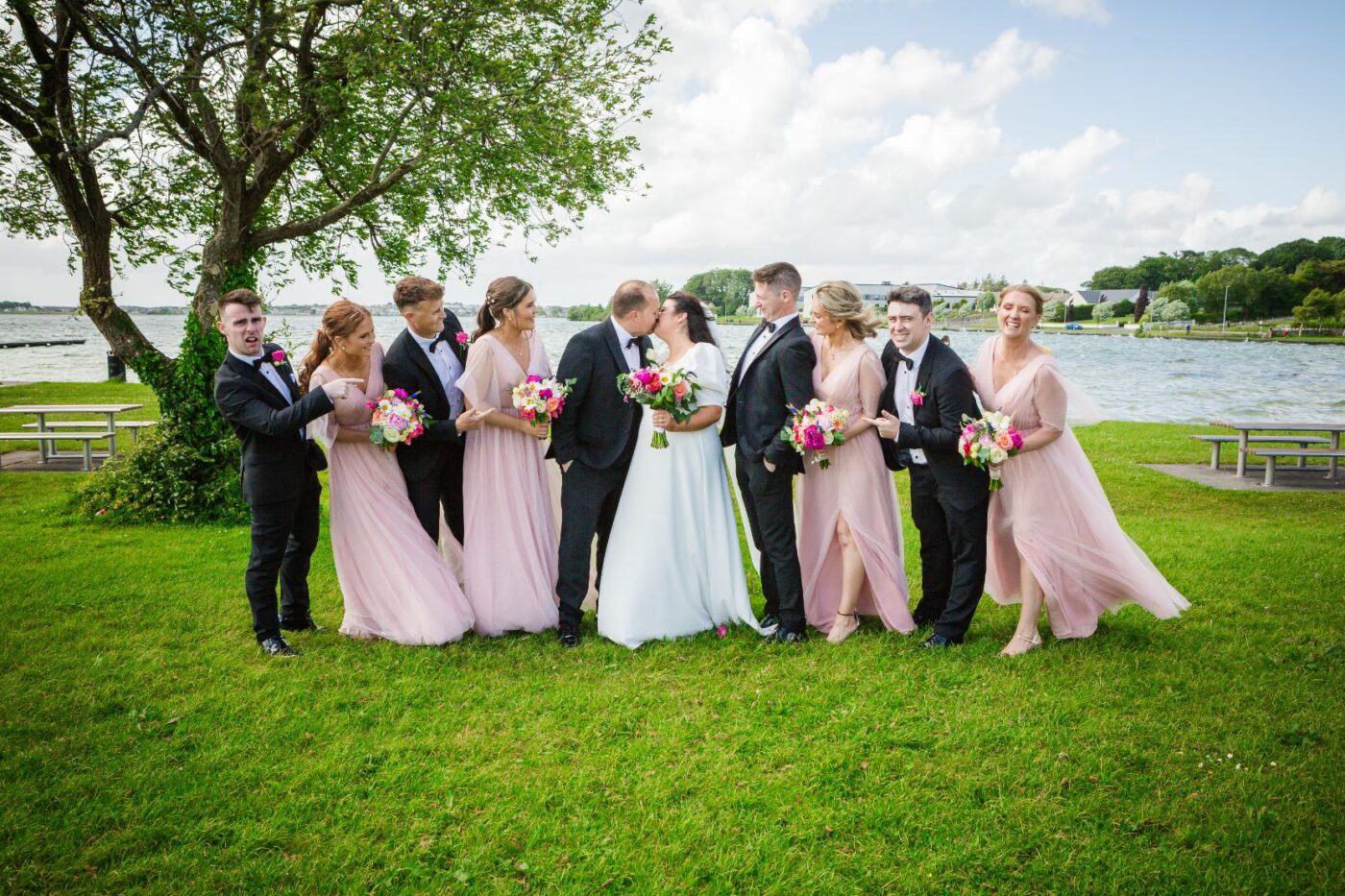 Bride and groom kissing beside Loughrea Lake as bridesmaids in blush pink and groomsmen in black laugh and point towards them.