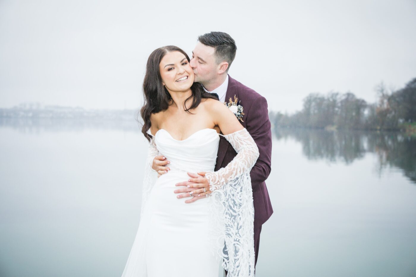 Groom in a burgundy suit embracing the smiling bride from behind and kissing her cheek beside a misty Loughrea Lake.