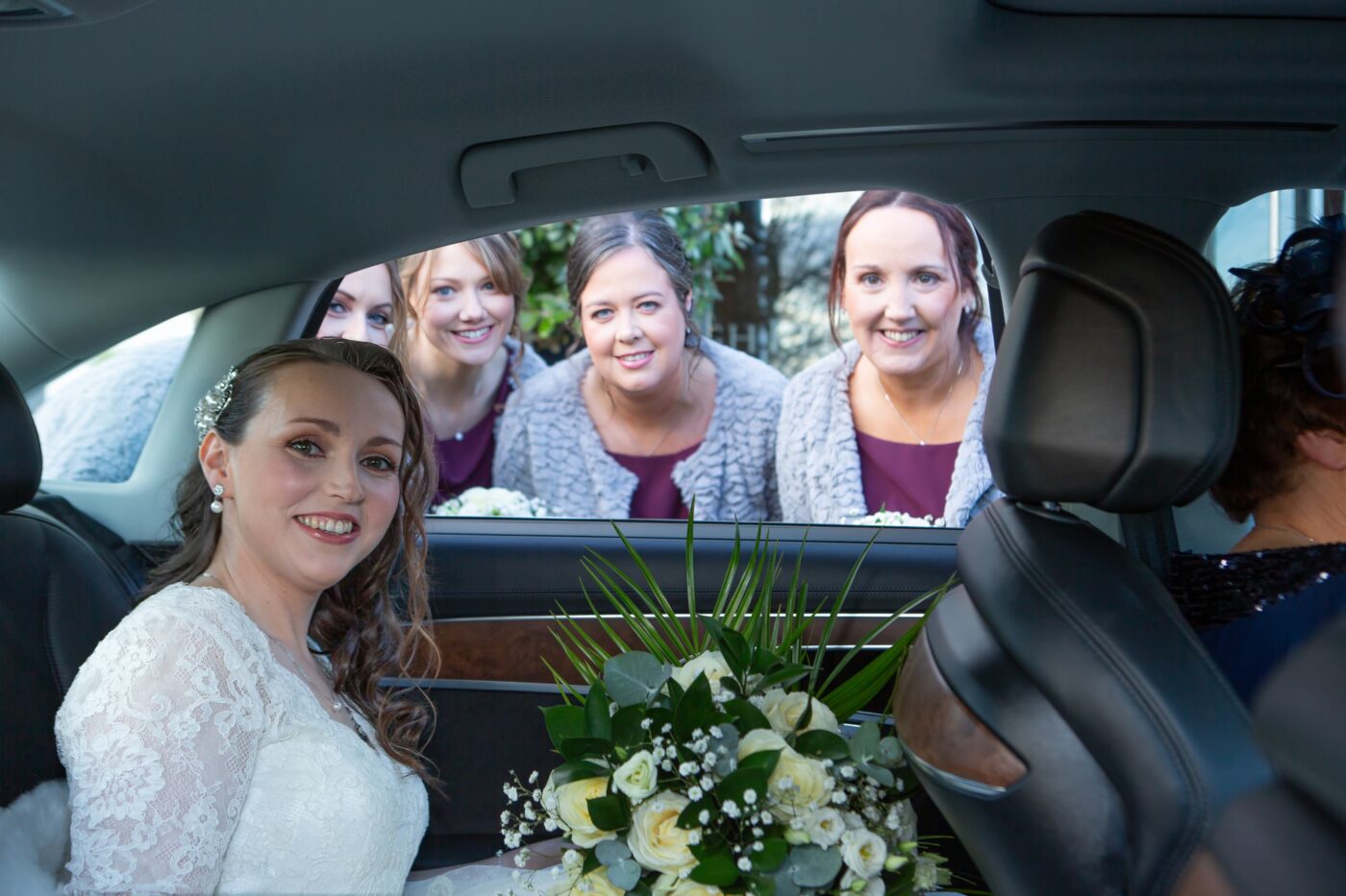 Bride sitting in her wedding car while four bridesmaids smile through the open window.