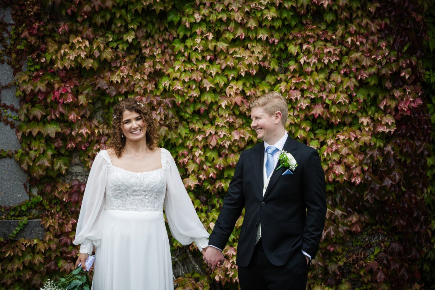Bride and groom smiling and holding hands in front of a wall covered in green and red ivy.