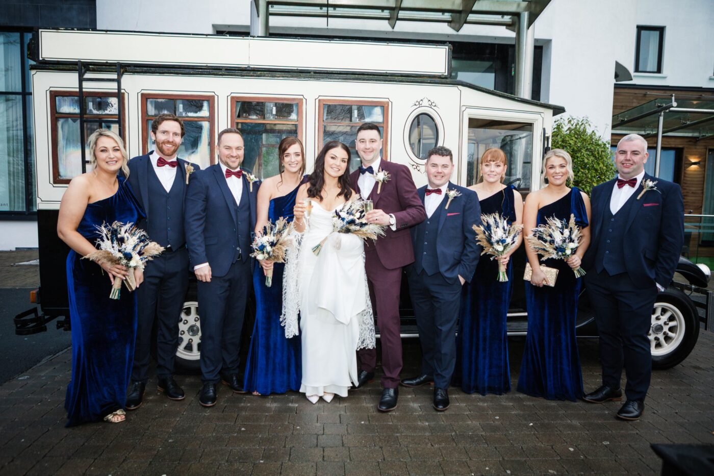 Bride and groom holding champagne glasses with their wedding party beside a cream vintage wedding bus.