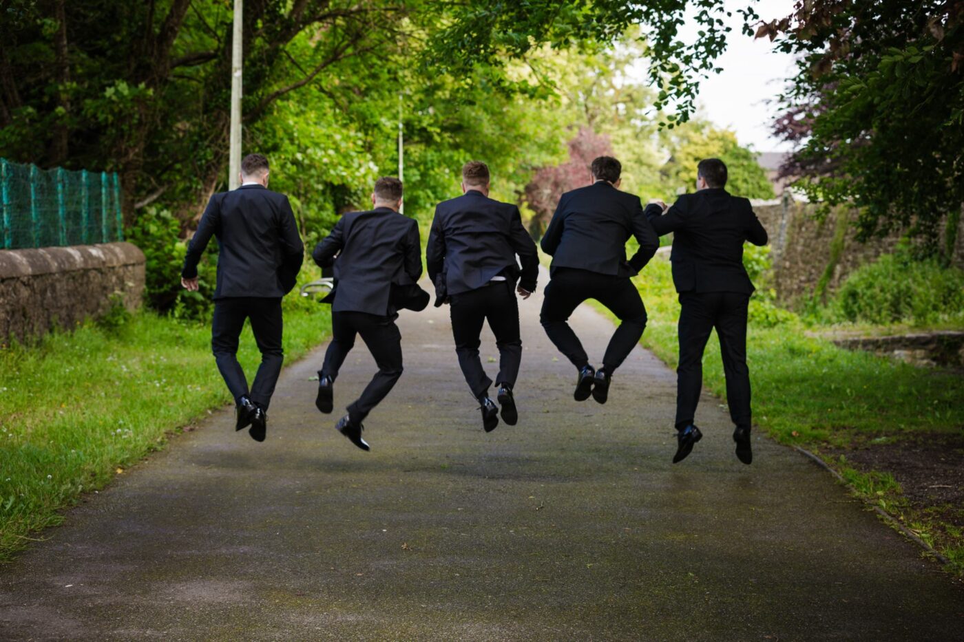 Five men in dark suits jumping with their backs to the camera on a tree-lined path in Loughrea.