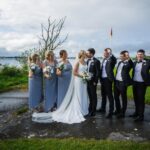 Áine and Rory kissing beside Loughrea Lake as their bridesmaids smile and groomsmen cheer.