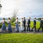 Áine and Rory standing together beside Loughrea Lake as their bridal party walks past in motion blur.