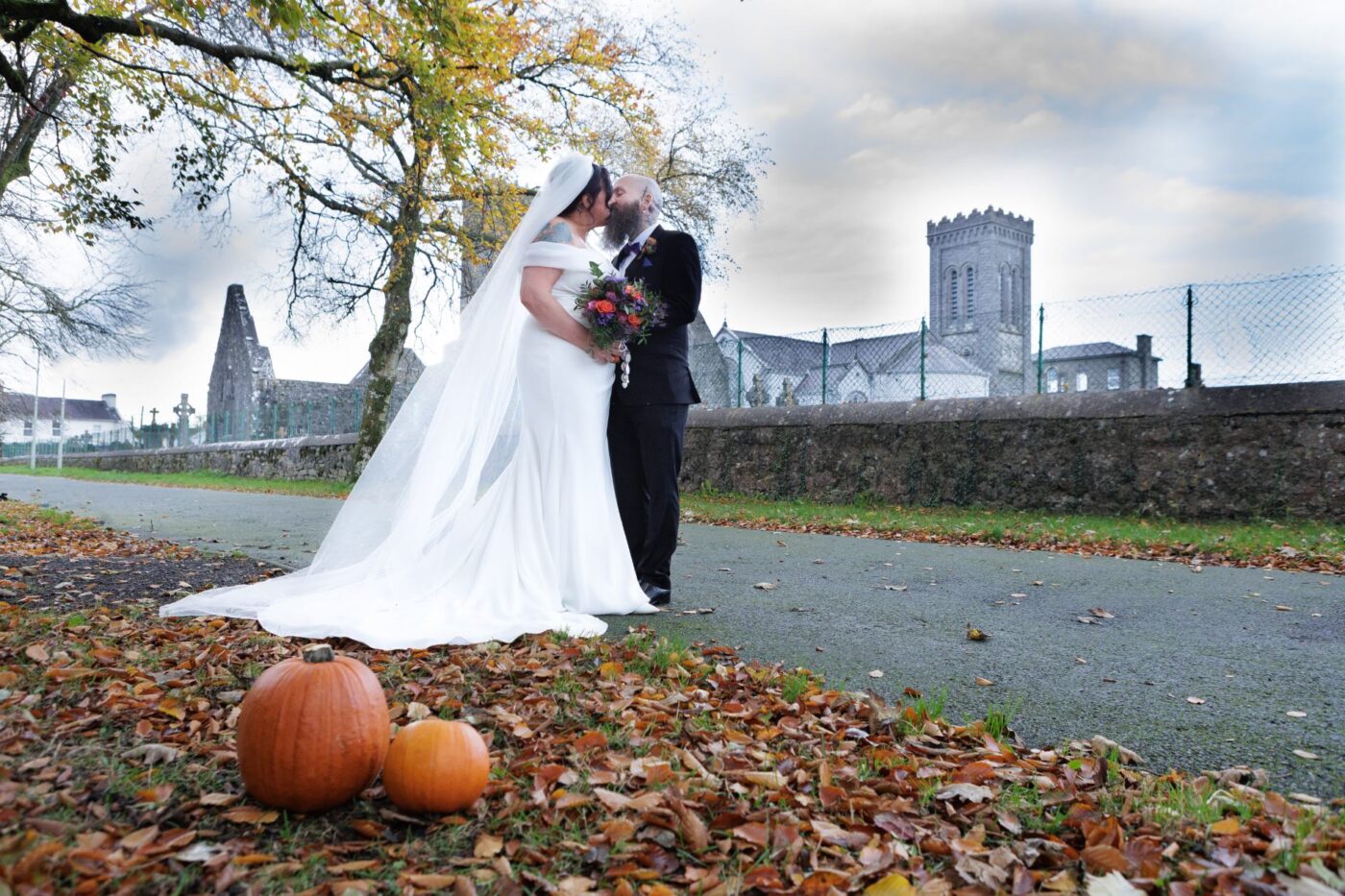 Bride and groom kissing beside stone abbey ruins, with orange pumpkins and fallen leaves in the foreground.