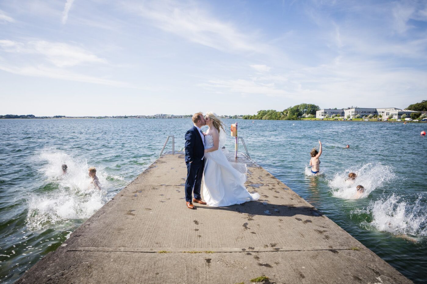 Bride and groom kissing on a pier at Loughrea Lake as swimmers jump into the water beneath a blue sky.