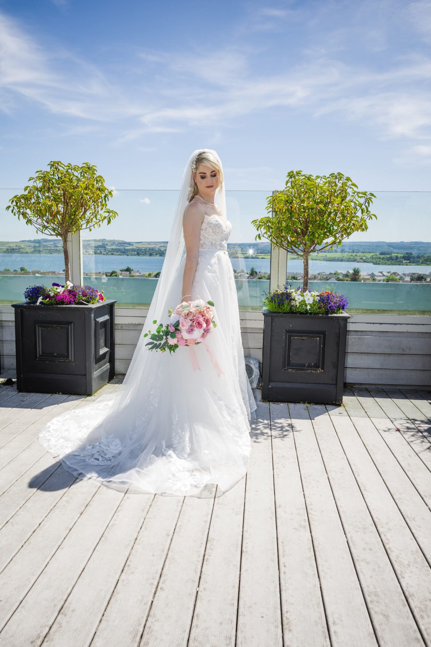 Bride holding a pink rose bouquet between potted trees on the sunny rooftop terrace at Loughrea Hotel & Spa, overlooking Loughrea Lake.