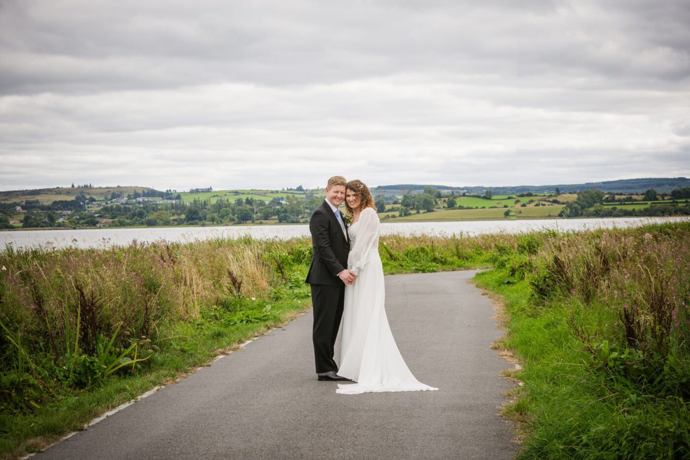 Bride and groom smiling and holding hands on a winding path beside Loughrea Lake, surrounded by grasses and wildflowers.
