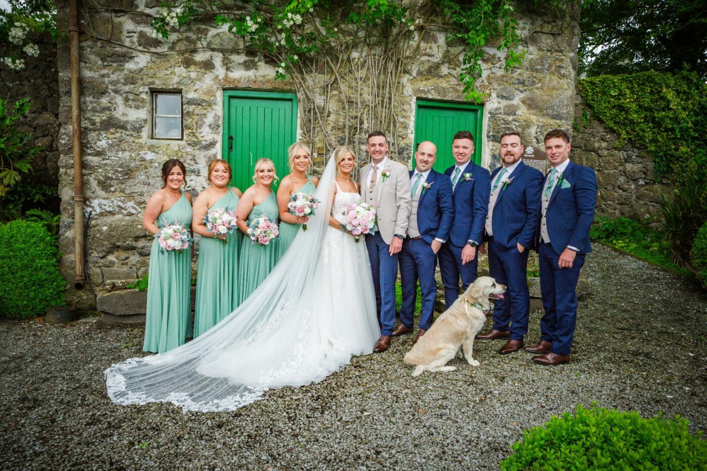 Bride and groom with bridesmaids in sage green, groomsmen in navy and a golden retriever outside a building with green doors.