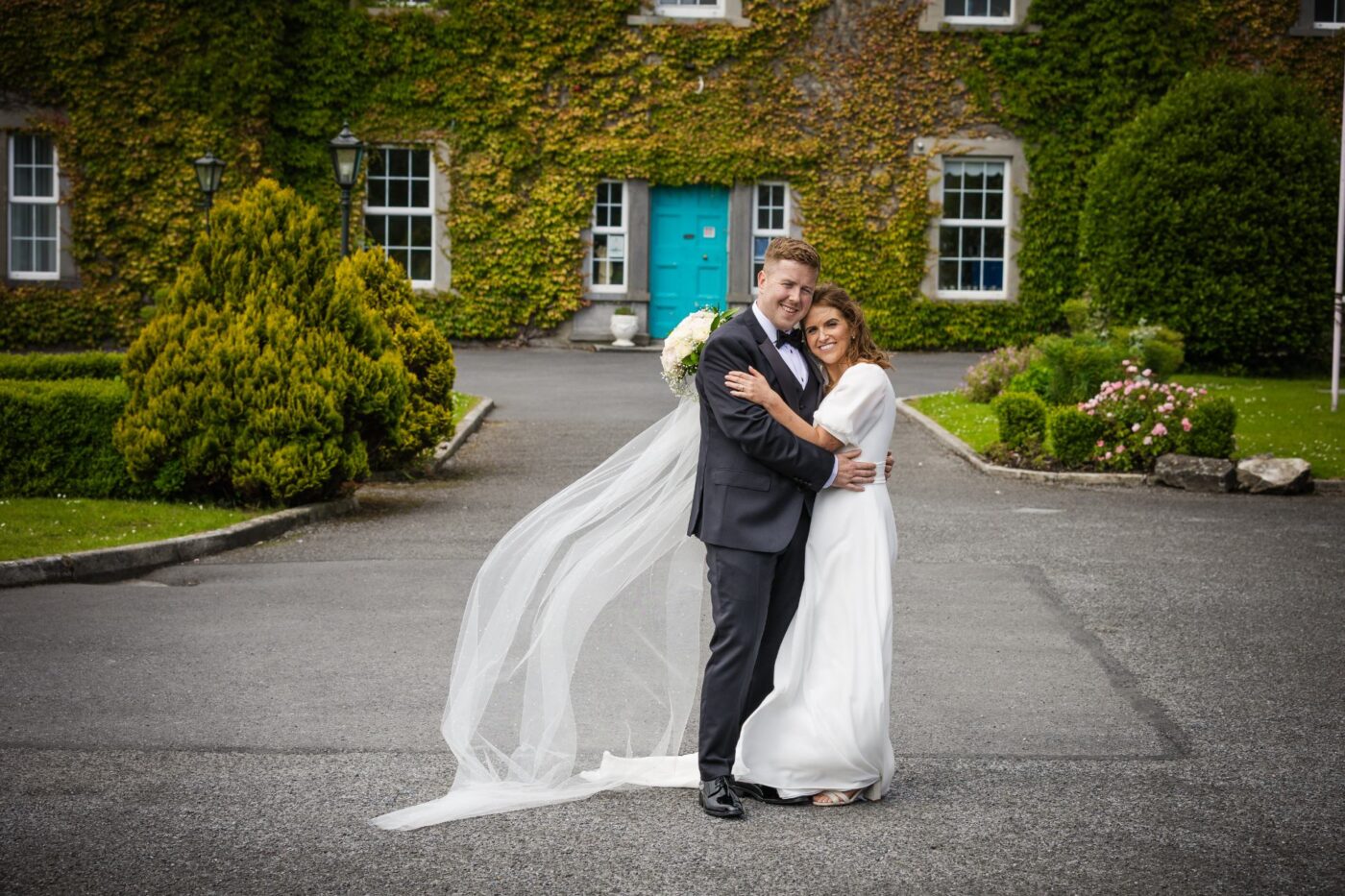 Bride and groom embracing outside an ivy-covered building with a turquoise door, with her veil flowing to the side.