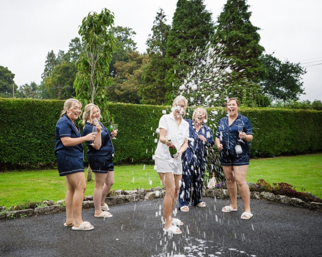 Áine spraying champagne outdoors as her mum and sisters laugh beside her.