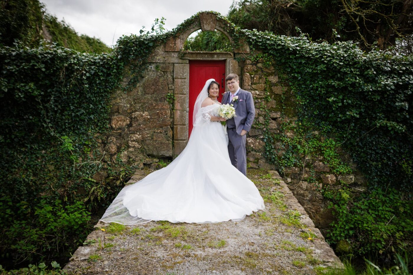 Bride and groom standing before a red door in an ivy-covered stone archway, with her dress train spread out.