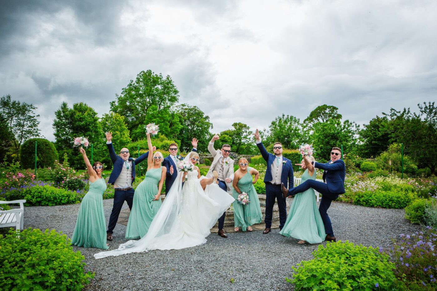 Bride, groom and wedding party wearing sunglasses and raising their arms in a colourful garden.