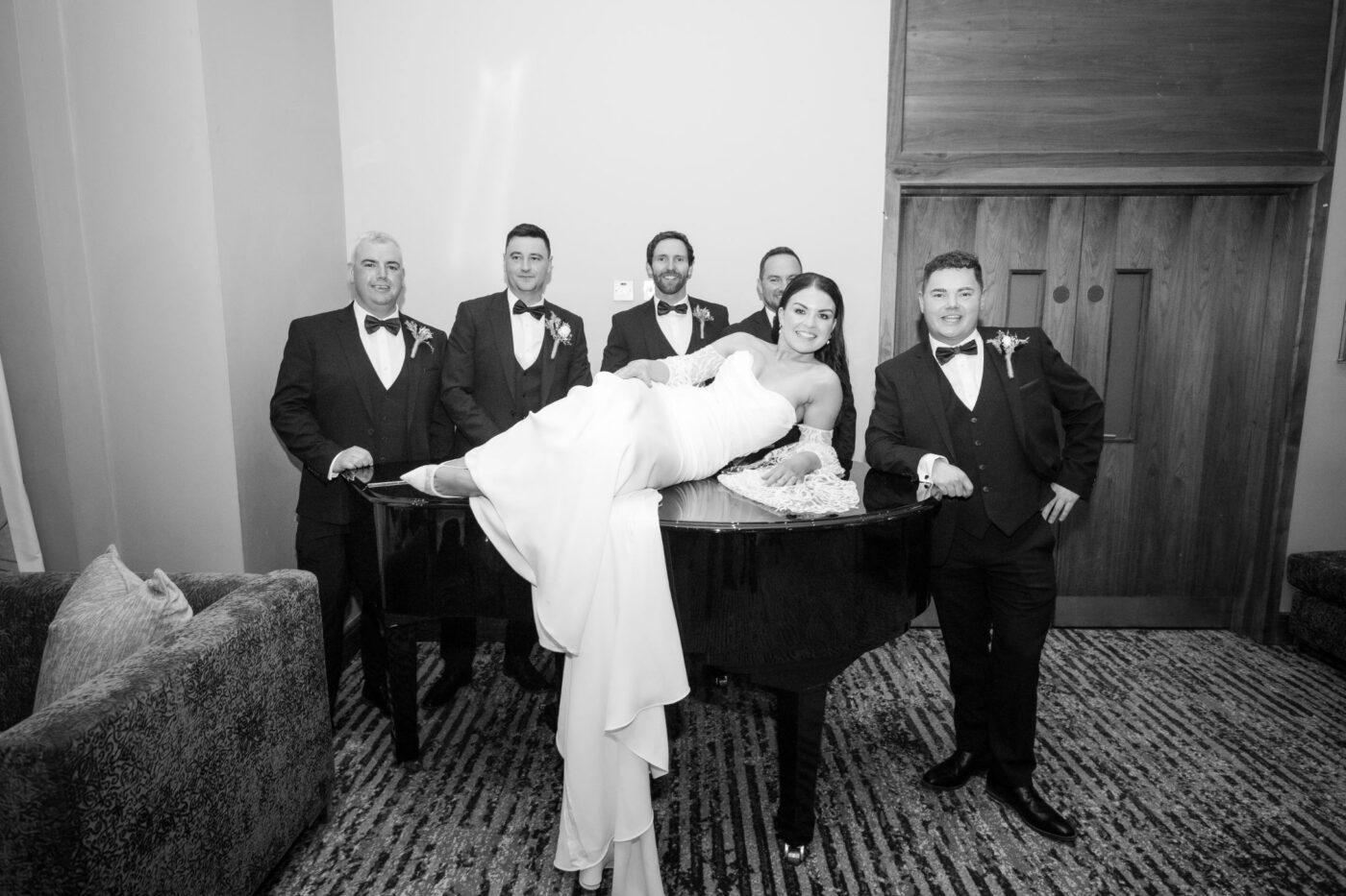 Black and white portrait of a bride reclining on a grand piano at Loughrea Hotel & Spa, surrounded by five men in black tuxedos.