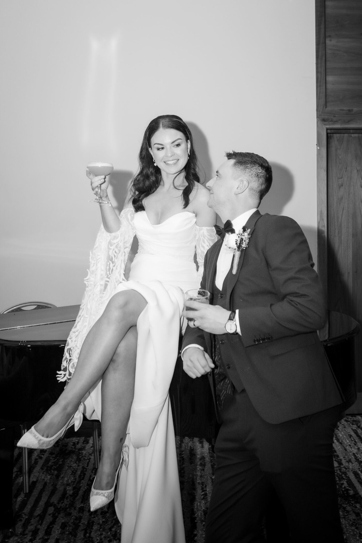 Black and white portrait of a bride sitting on a piano beside the groom at Loughrea Hotel & Spa, both holding drinks and smiling at each other.