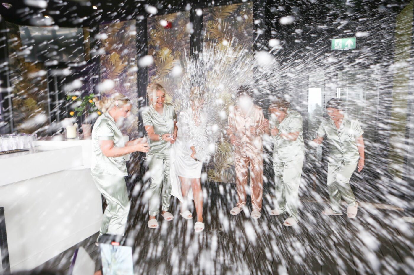 Bride and friends in satin nightwear laughing as champagne sprays across the room.