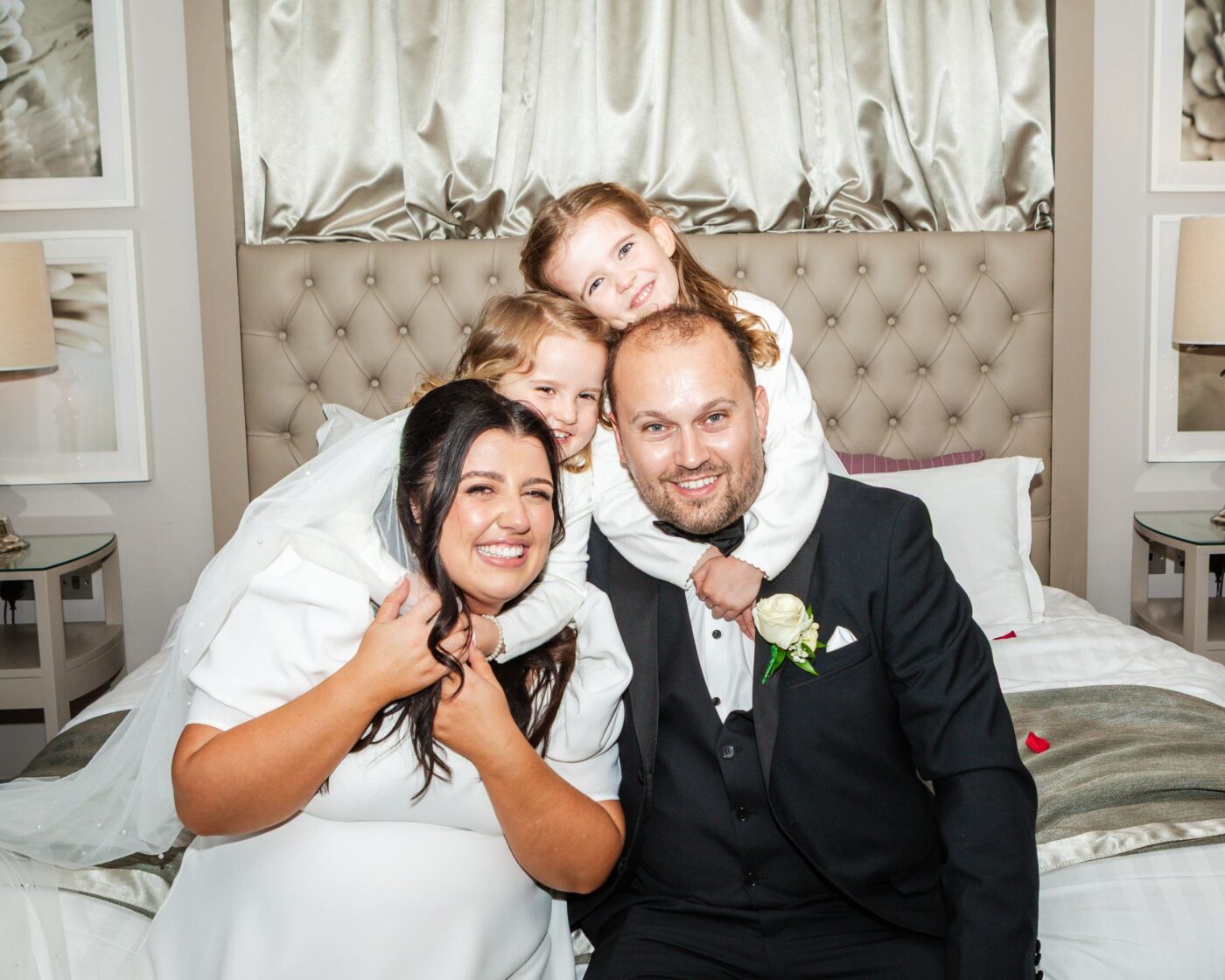 Bride and groom smiling on a bed as two young girls hug them from behind.