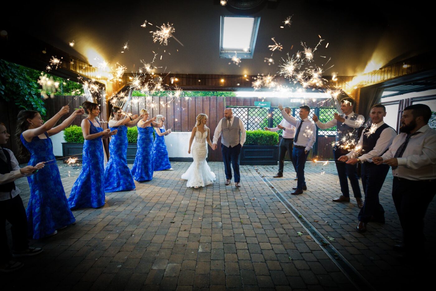 Bride and groom walking hand in hand between bridesmaids in blue floral dresses and groomsmen holding sparklers at Loughrea Hotel & Spa.