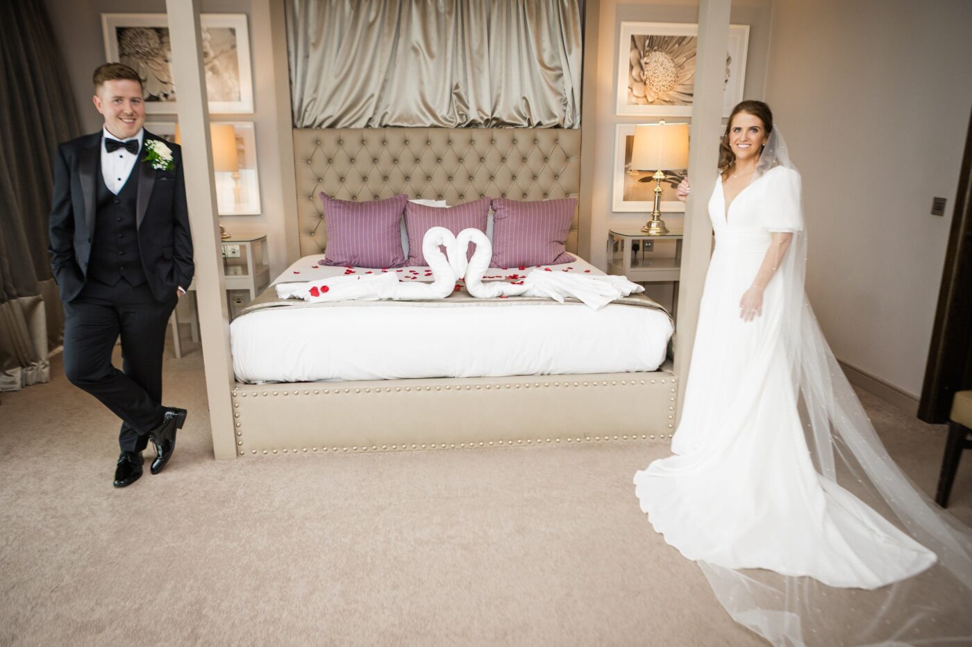 Bride and groom standing on either side of a four-poster bed decorated with towel swans and rose petals in the bridal suite at Loughrea Hotel & Spa.