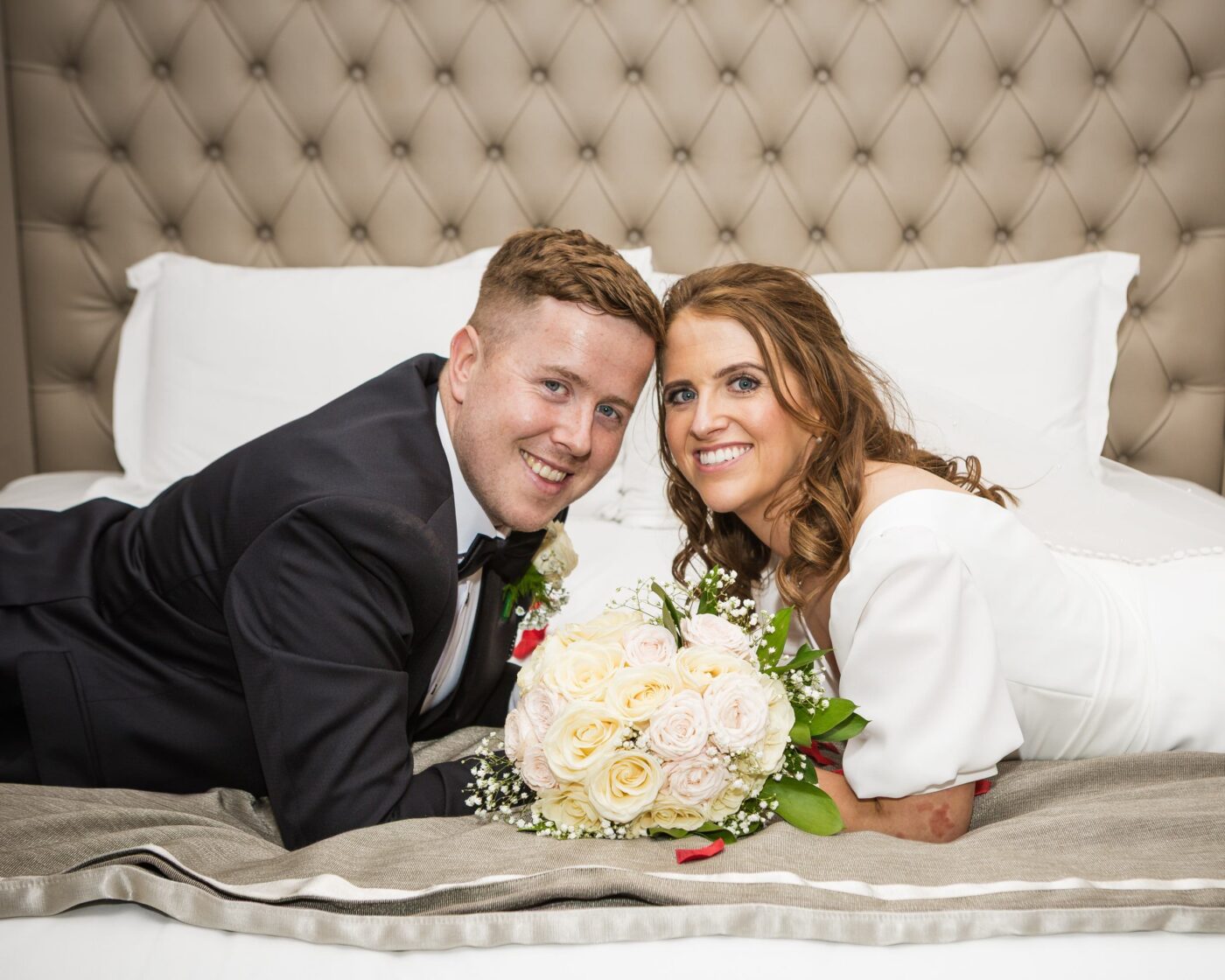 Bride and groom lying together on a bed at Loughrea Hotel & Spa, smiling beside a bouquet of cream and pale pink roses.