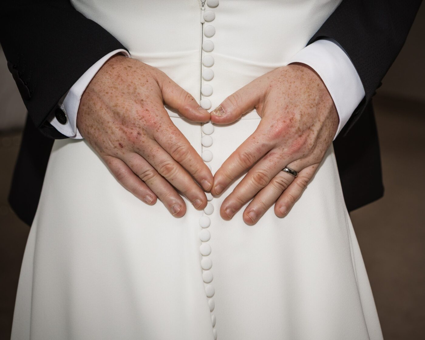 Close-up of the groom’s hands forming a heart against the buttons of the bride’s white wedding