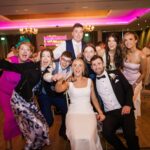 Áine and Rory laughing and posing with cheering wedding guests beneath pink lighting at Loughrea Hotel & Spa.