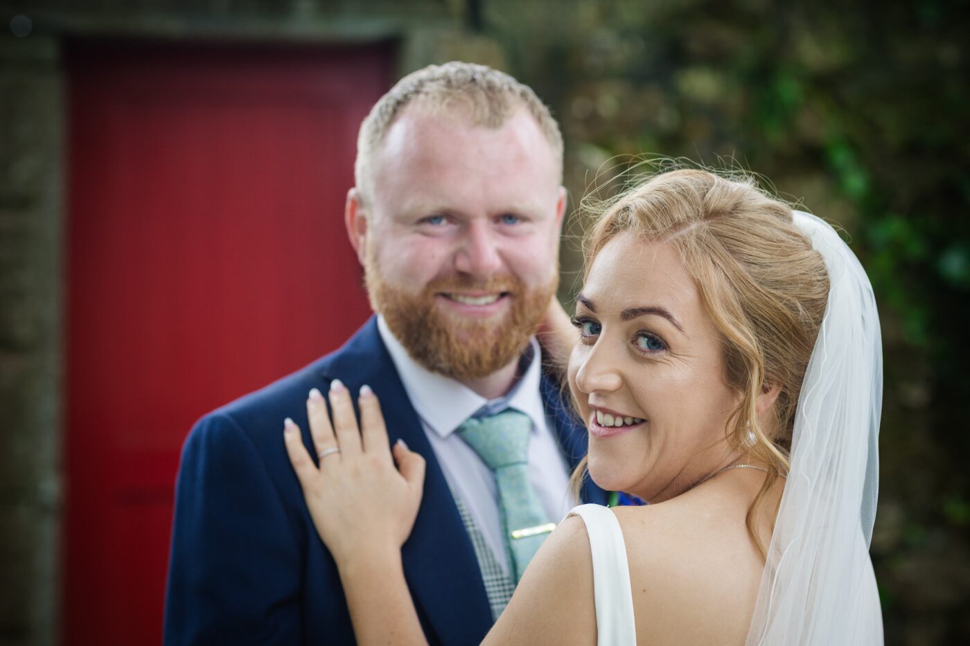 Smiling bride resting her hand on the groom’s shoulder, with a red door behind them.