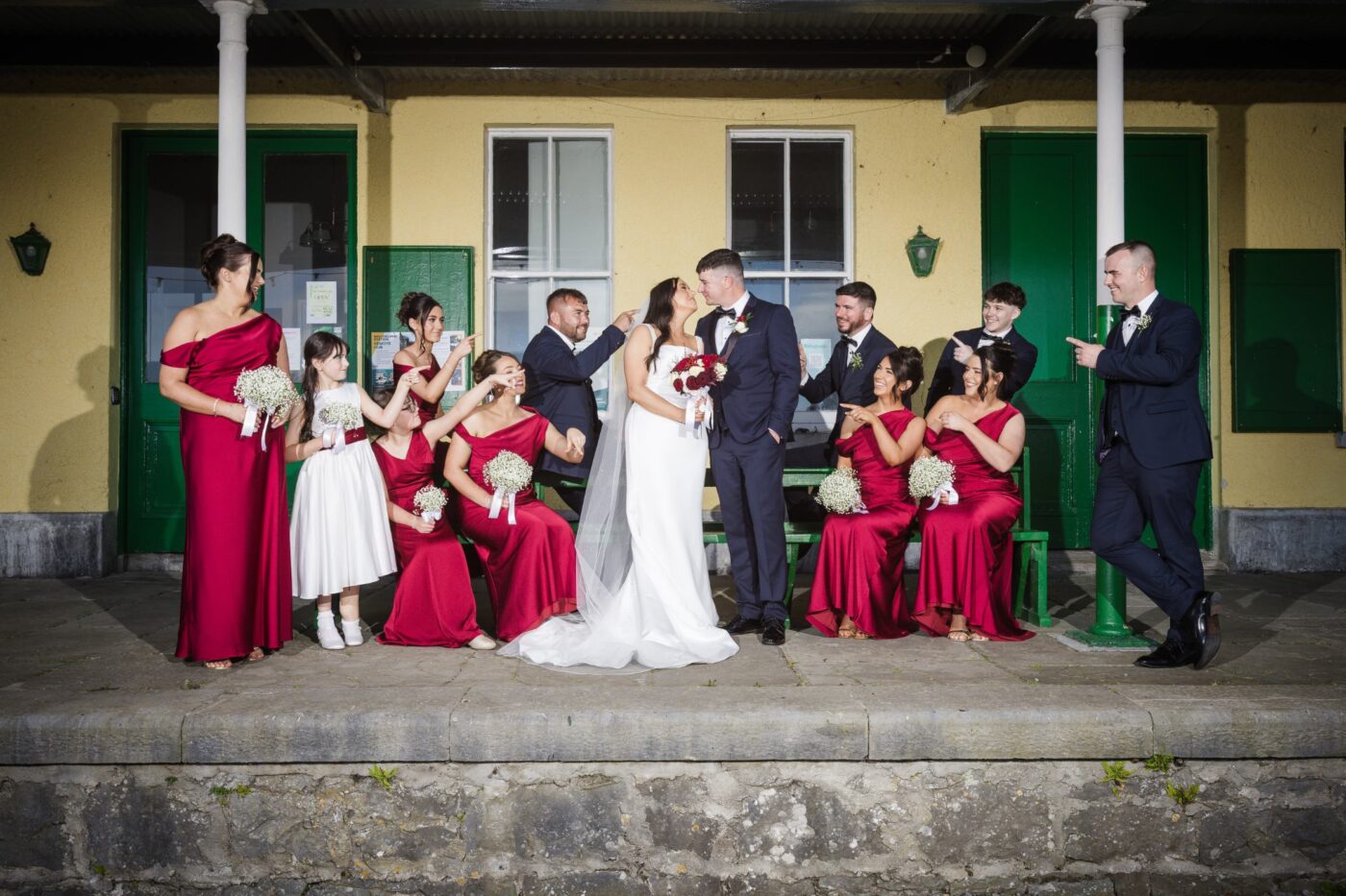 Bride and groom kissing on a railway platform as bridesmaids in burgundy and groomsmen point towards them.