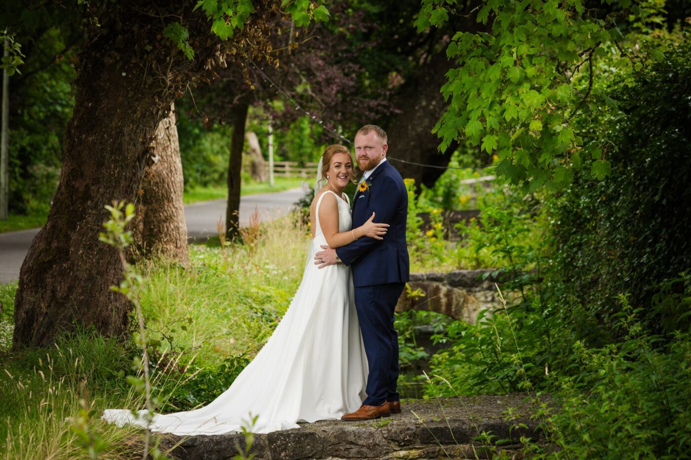 Bride and groom embracing on a stone bridge beneath leafy trees beside a stream.