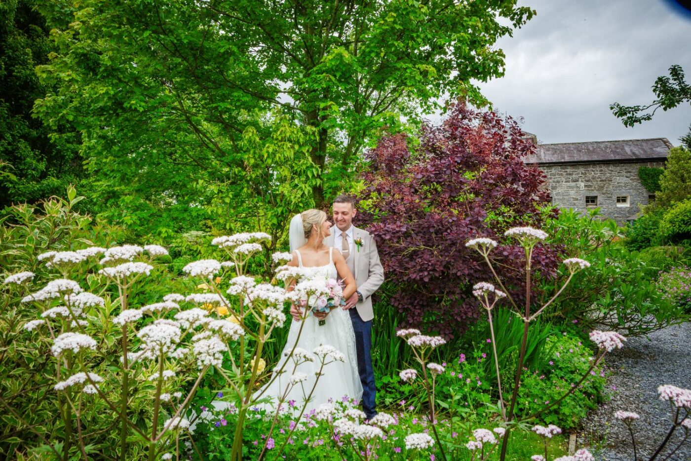 Bride and groom embracing in a lush garden, framed by tall white flowers and leafy trees.