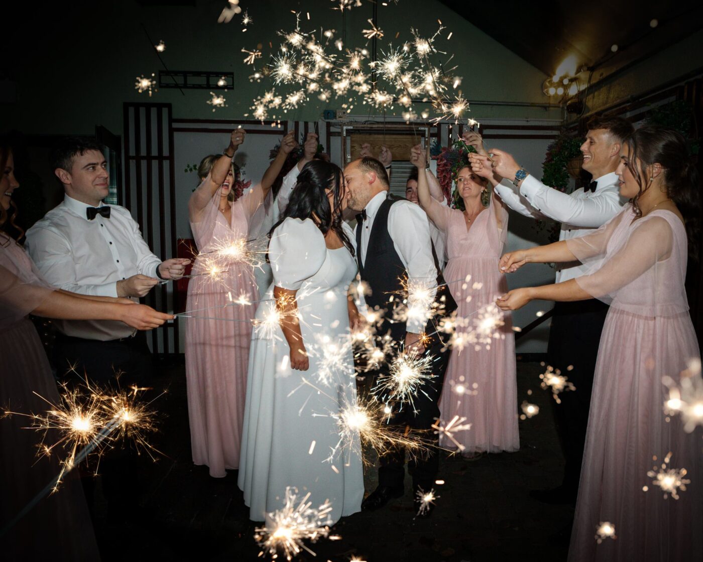 Bride and groom kissing at Loughrea Hotel & Spa as bridesmaids in blush pink and groomsmen surround them with glowing sparklers.