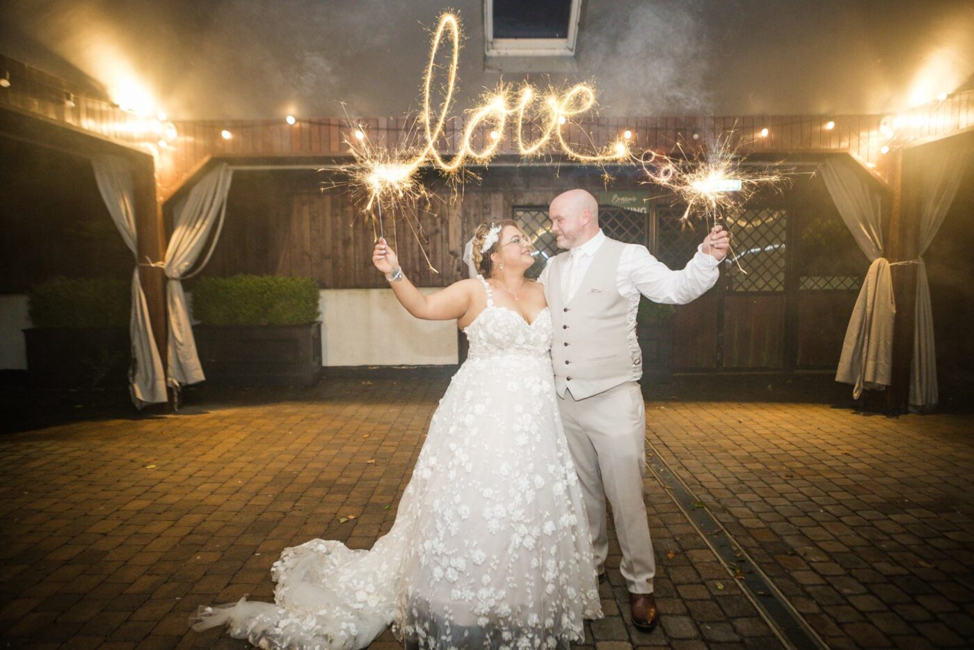 Bride and groom holding sparklers beneath a glowing light trail spelling “love”.