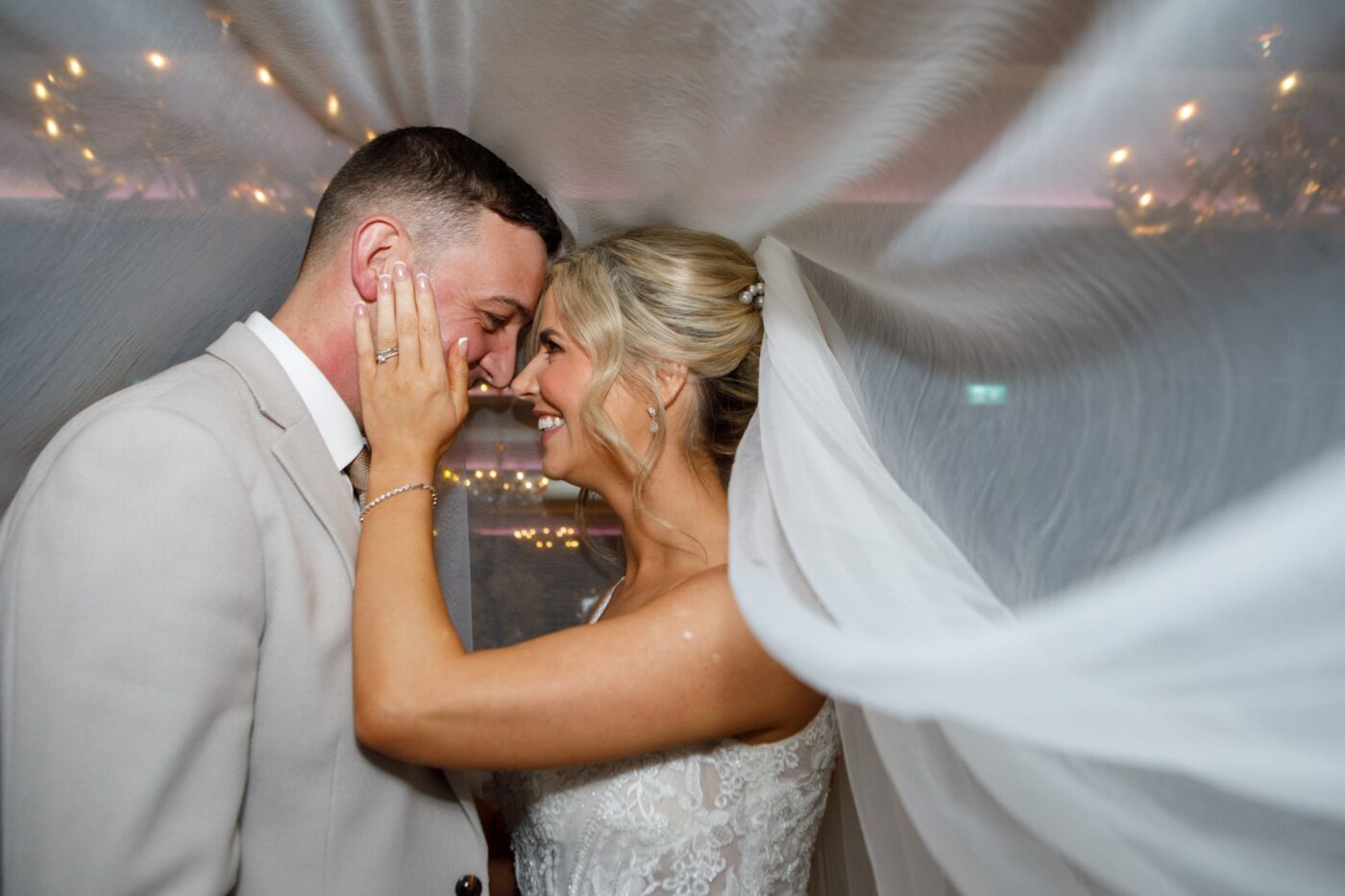Bride and groom smiling with their foreheads together beneath her flowing veil, with warm lights behind them.