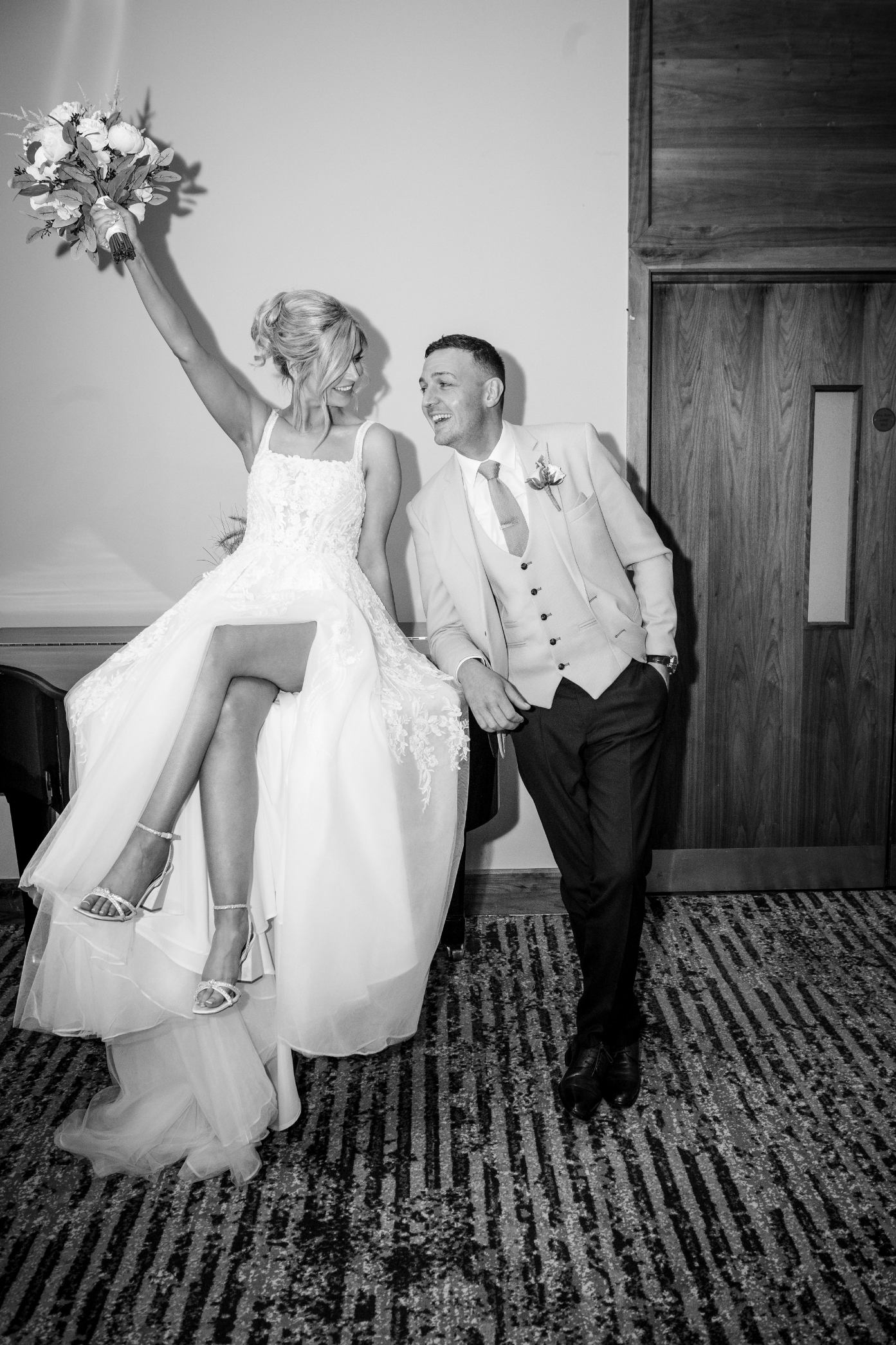 Black and white portrait of a bride sitting on a piano and raising her bouquet beside the smiling groom at Loughrea Hotel & Spa.