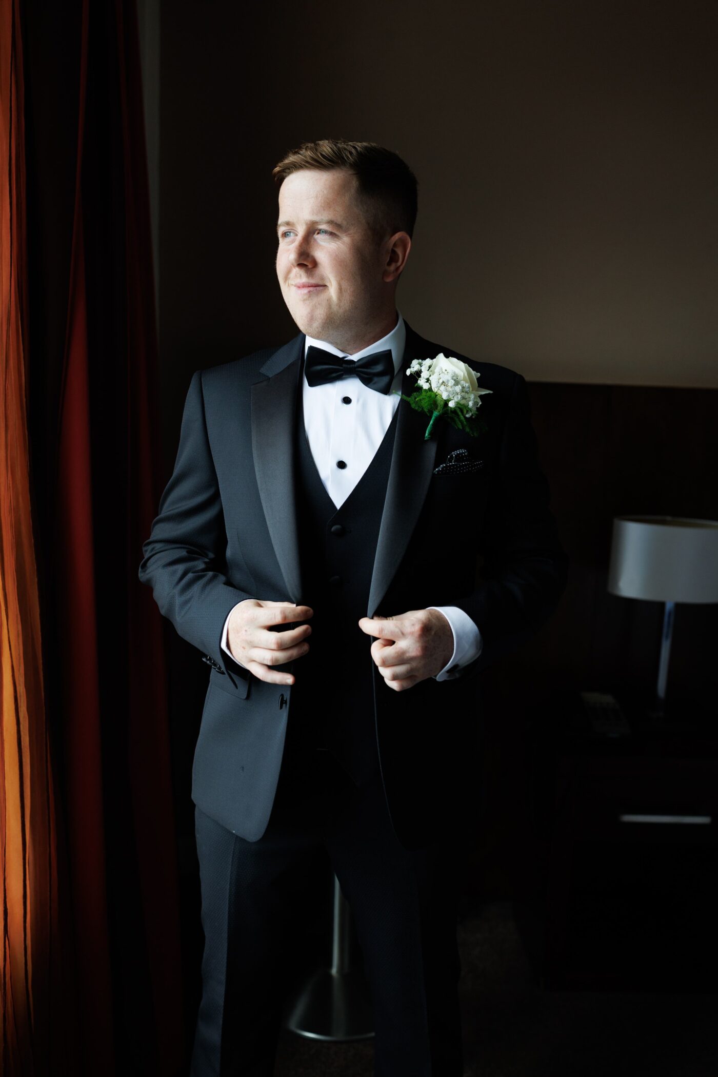 Groom wearing a black tuxedo, bow tie and white boutonnière, looking towards the window while holding his jacket.