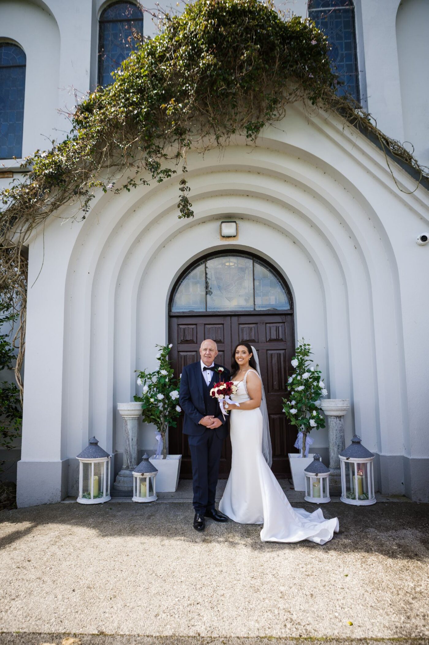 Bride holding a burgundy and white bouquet beside her dad in a navy suit at a church entrance decorated with flowers and lanterns.