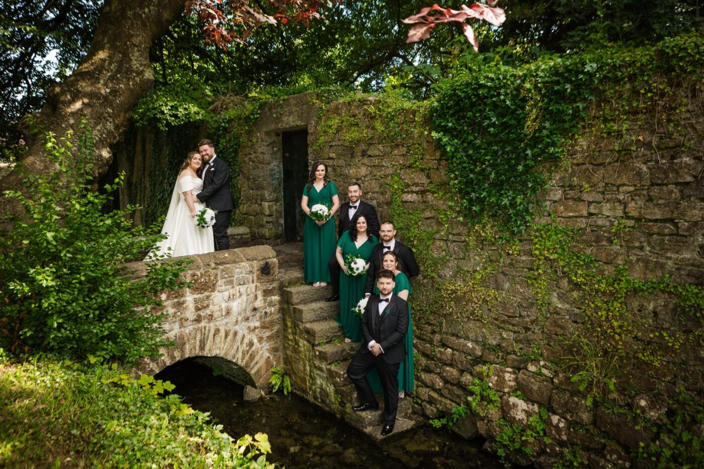 Bride and groom with bridesmaids in emerald green and groomsmen in black on stone steps beside an ivy-covered wall and stream.