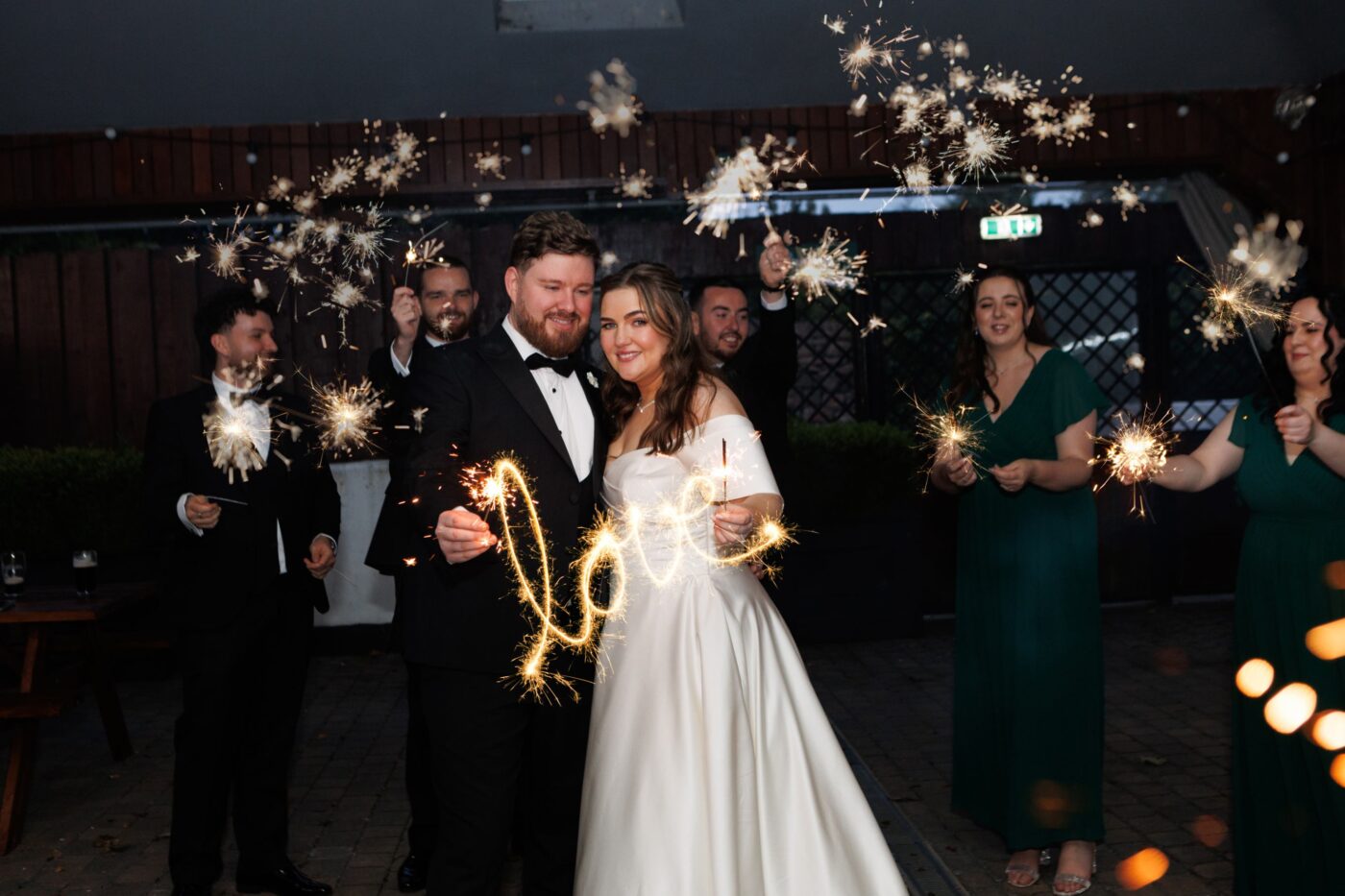 Bride and groom holding sparklers with a light trail spelling “love”, surrounded by their wedding party.