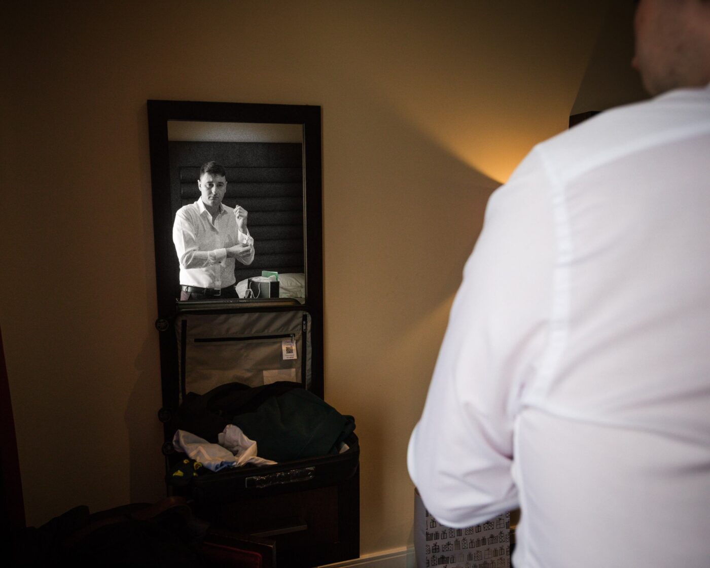 Groom reflected in a bedroom mirror as he adjusts the cuff of his white shirt.