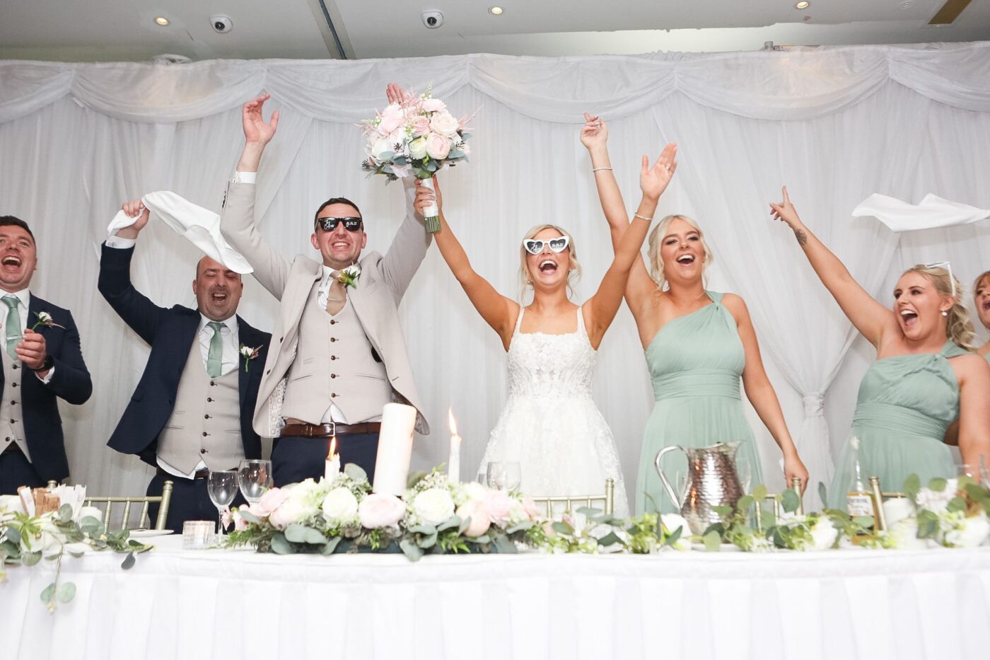 Bride, groom and wedding party cheering with raised arms behind a table decorated with flowers and candles at Loughrea Hotel & Spa.