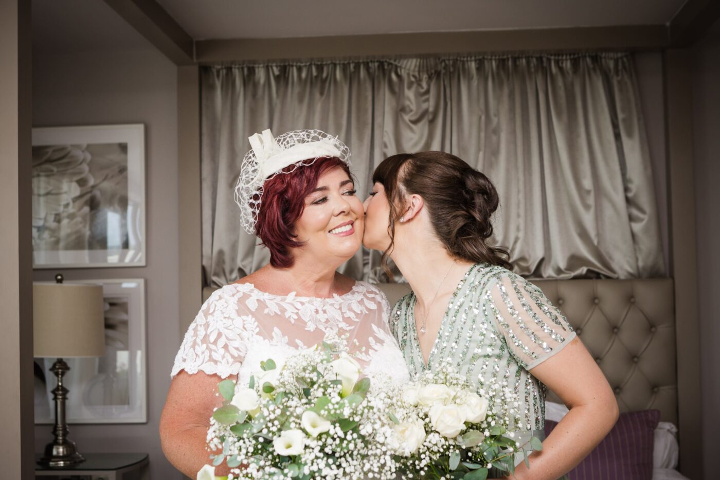Woman in a green sequinned dress kissing the cheek of a smiling bride holding a white bouquet.