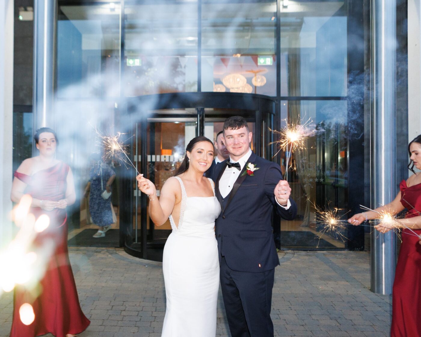 Bride and groom holding lit sparklers outside a glass hotel entrance, surrounded by bridesmaids in burgundy.