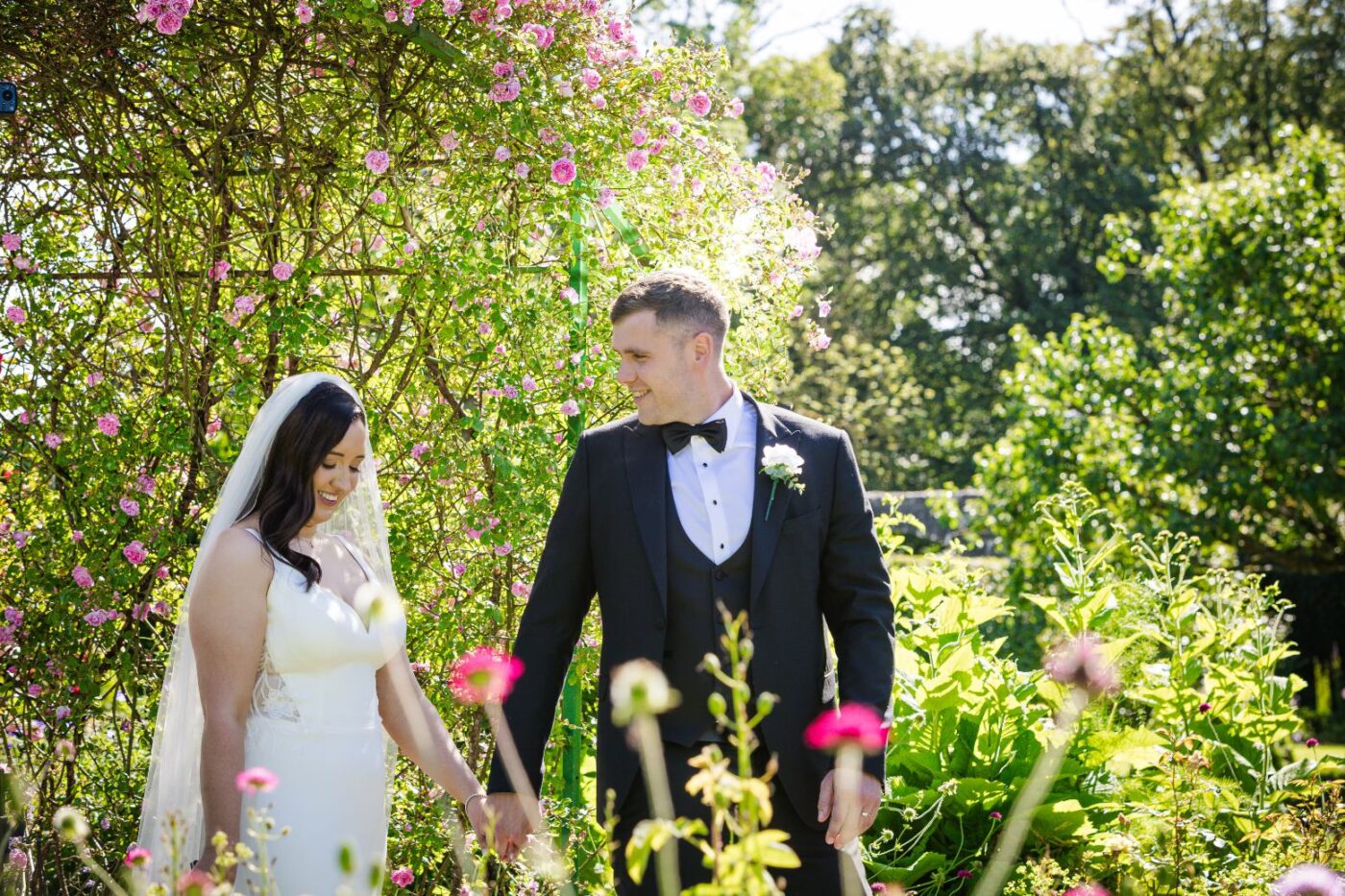 Bride and groom smiling and holding hands in a sunlit garden beneath pink climbing roses.