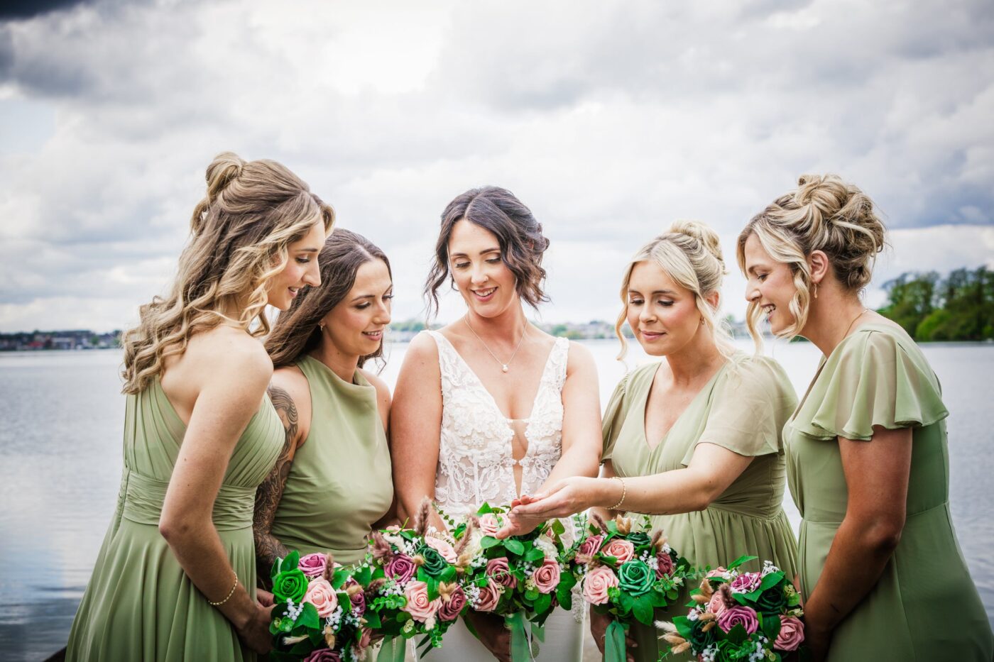 Bride and four bridesmaids in sage green looking at her wedding ring beside Loughrea Lake, holding colourful bouquets.
