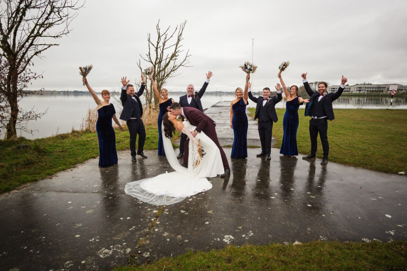 Groom dipping and kissing the bride as their wedding party raises bouquets and cheers beside Loughrea Lake.