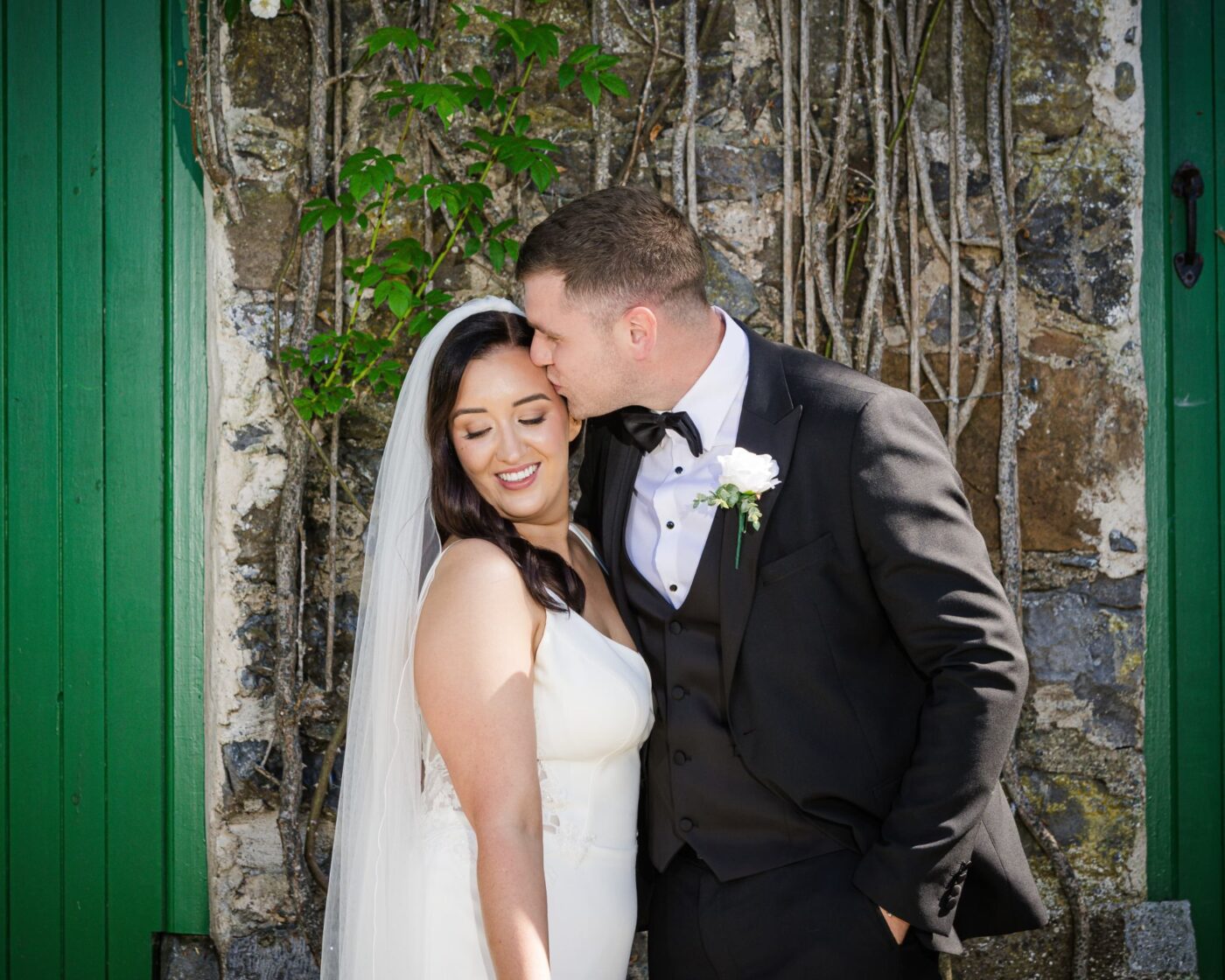 Groom kissing the smiling bride’s forehead beside a stone wall and green doors.