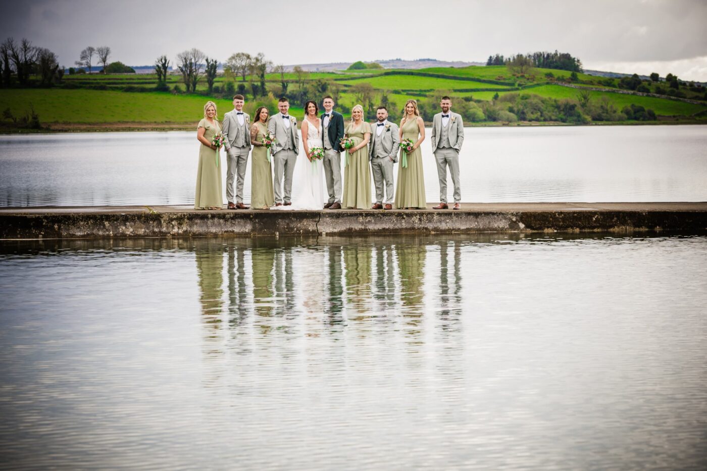 Bride and groom with bridesmaids in sage green and groomsmen in grey standing on a pier, reflected in Loughrea Lake.