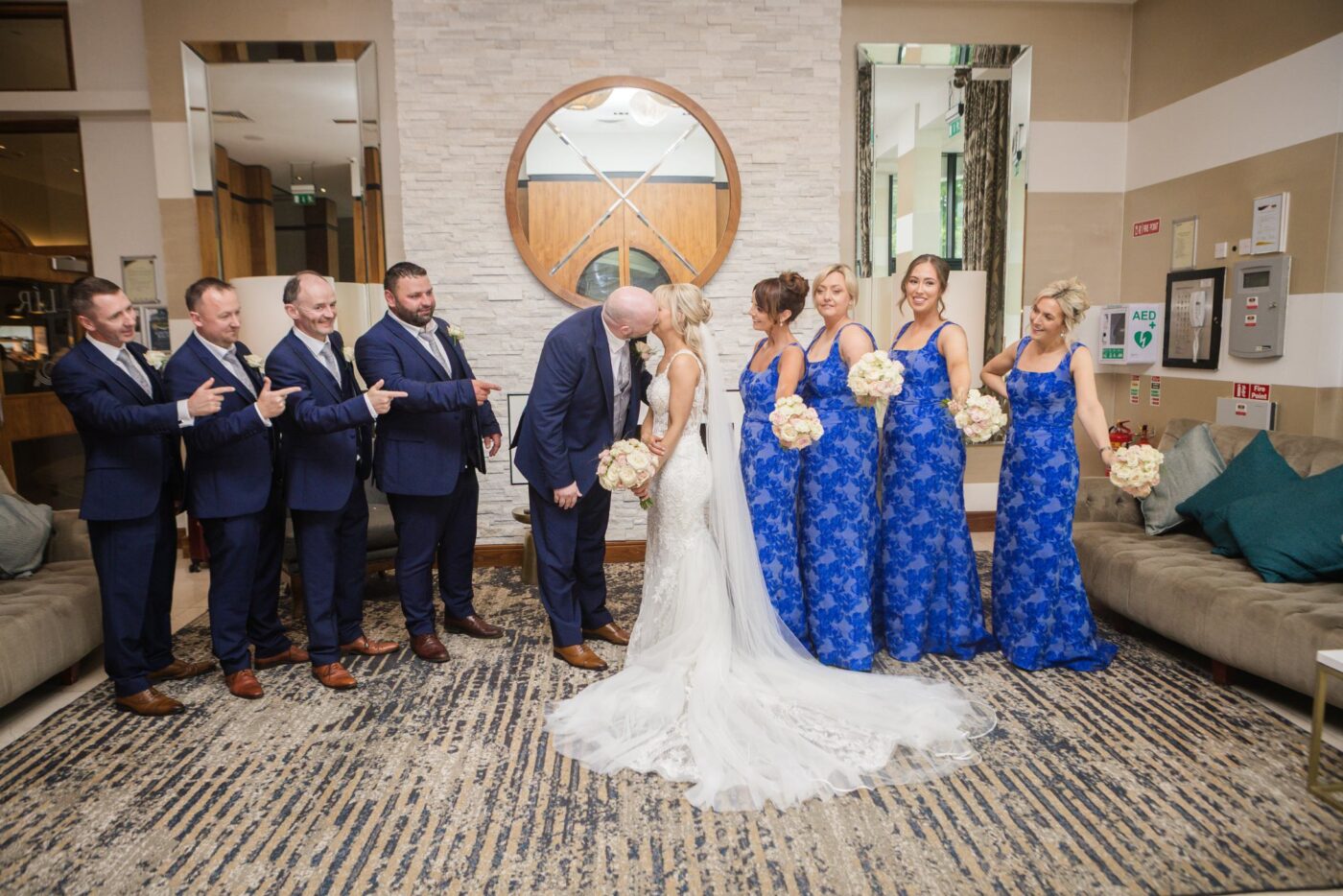 Bride and groom kissing beside a fireplace at Loughrea Hotel & Spa, flanked by bridesmaids in blue floral dresses and groomsmen in navy.